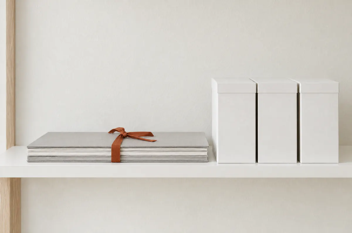 Illustration of a closed, bound drawing set tied with a rust-coloured ribbon on a white shelf beside archive boxes.